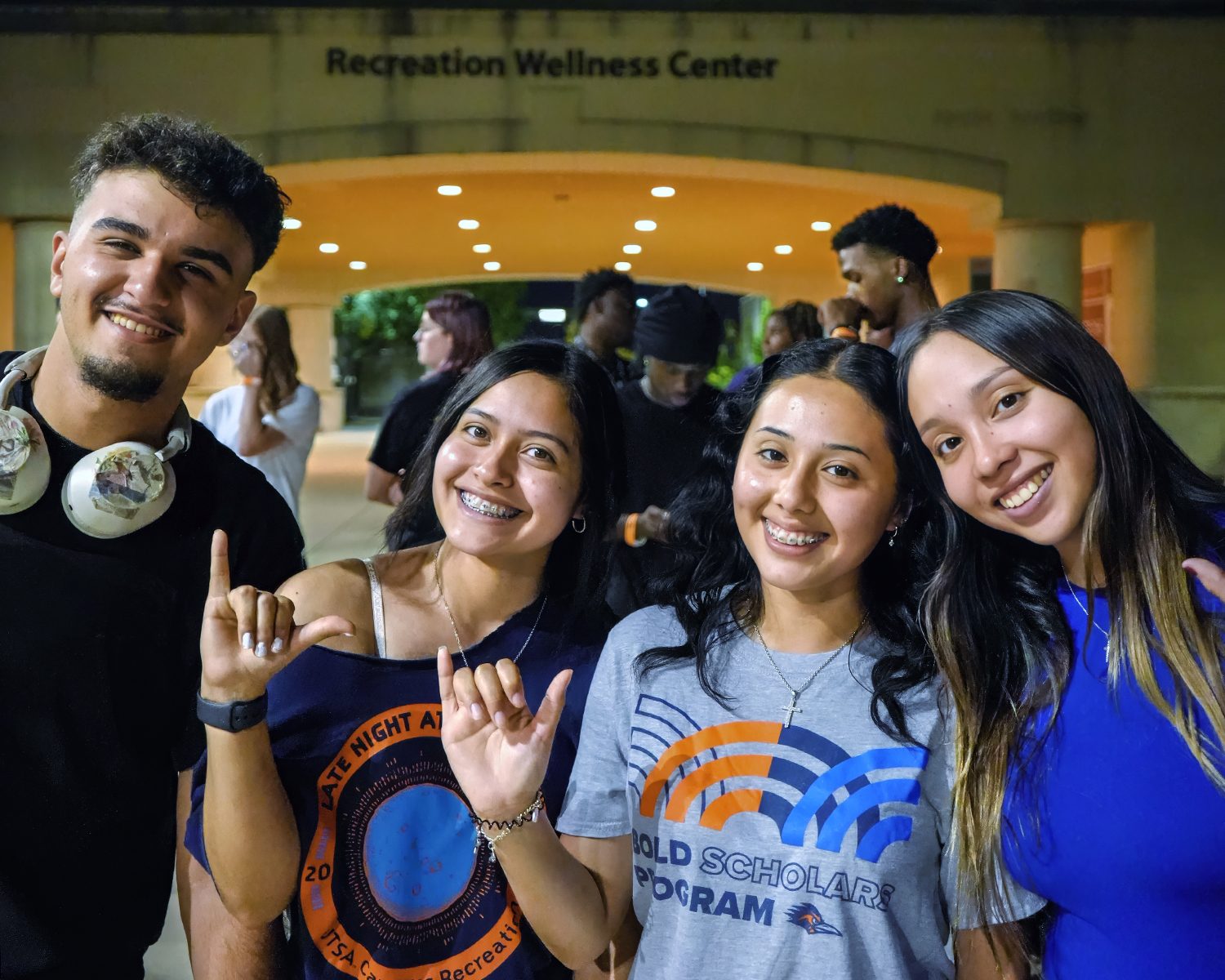 From left to right, a group of students posing, a man with headphones smiling, three women smiling and holding up the school "birds up" hand gesture.